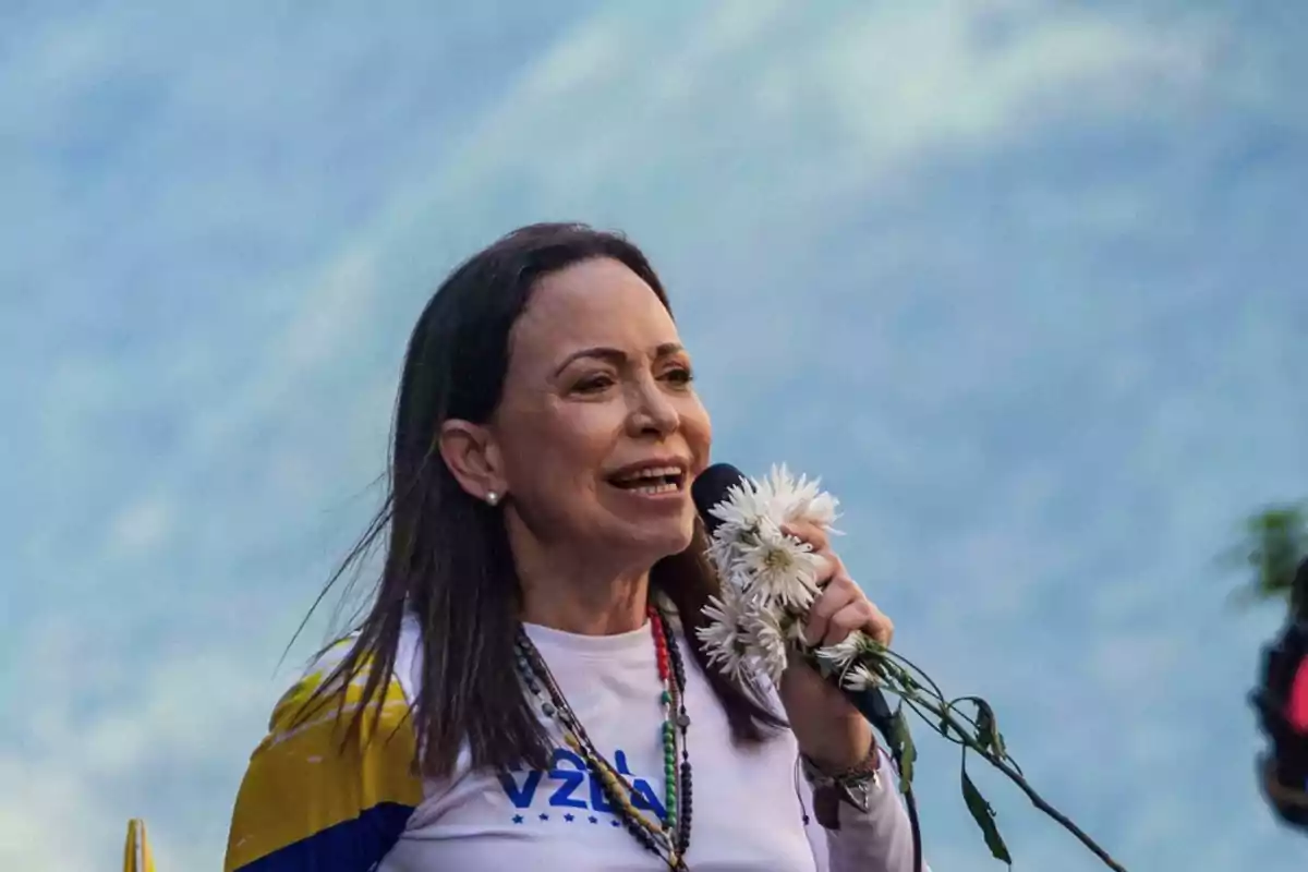 Mujer de cabello oscuro hablando por un micrófono y sosteniendo flores blancas, lleva puesta una camiseta blanca con letras azules y una bandera sobre los hombros, el fondo es desenfocado y azul. Mujer de cabello oscuro hablando por un micrófono y sosteniendo flores blancas, lleva puesta una camiseta blanca con letras azules y una bandera sobre los hombros, el fondo es desenfocado y azul.