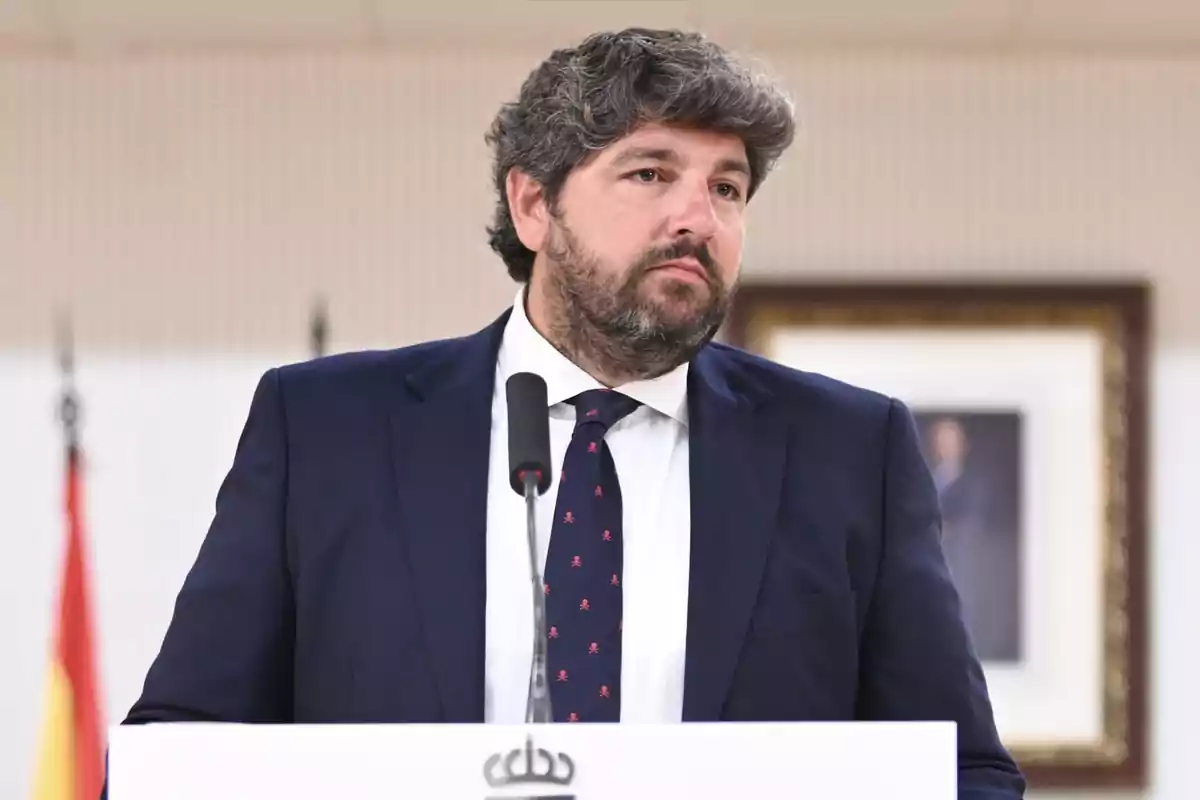 Hombre de cabello rizado y barba con traje azul y corbata hablando frente a un micrófono en un podio, con una bandera y un cuadro desenfocados al fondo