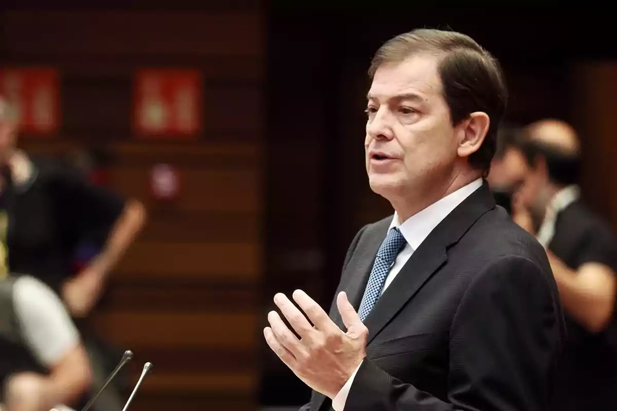 Hombre de traje y corbata hablando en público en un entorno formal con personas desenfocadas al fondo Hombre de traje y corbata hablando en público en un entorno formal con personas desenfocadas al fondo