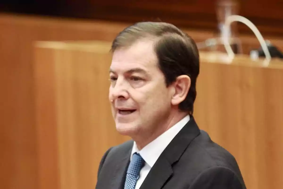 Hombre de mediana edad con cabello oscuro y traje formal hablando en un entorno institucional con fondo de madera