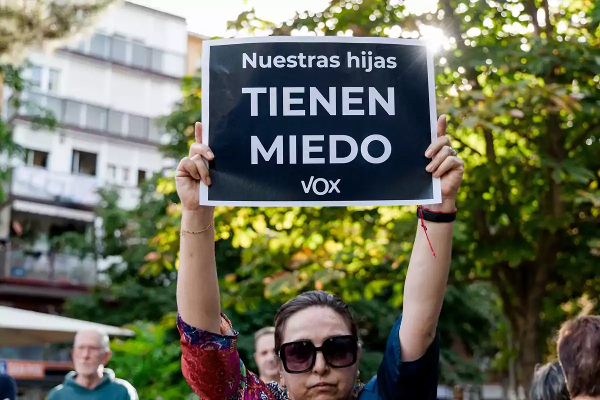 Mujer con gafas de sol sostiene un cartel negro con letras blancas que dice Nuestras hijas tienen miedo VOX durante una manifestación al aire libre Mujer con gafas de sol sostiene un cartel negro con letras blancas que dice Nuestras hijas tienen miedo VOX durante una manifestación al aire libre