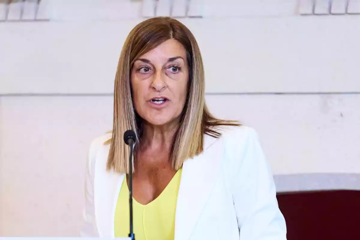 Mujer de cabello lacio y rubio oscuro usando saco blanco y blusa amarilla hablando frente a un micrófono Mujer de cabello lacio y rubio oscuro usando saco blanco y blusa amarilla hablando frente a un micrófono