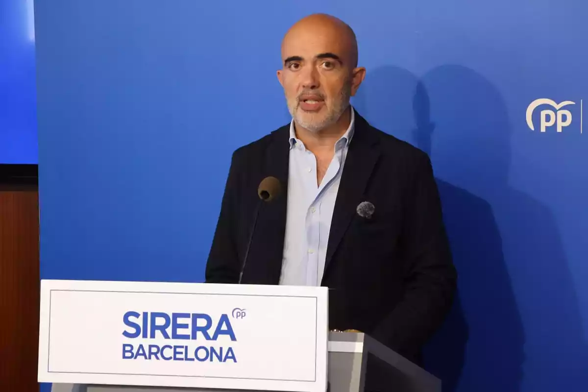 Hombre calvo con barba hablando en un atril con el nombre Sirera Barcelona y fondo azul con el logo del Partido Popular Hombre calvo con barba hablando en un atril con el nombre Sirera Barcelona y fondo azul con el logo del Partido Popular