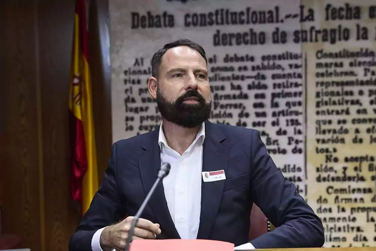 Hombre con barba y traje oscuro sentado frente a un micrófono en una sala con fondo de texto y una bandera de España