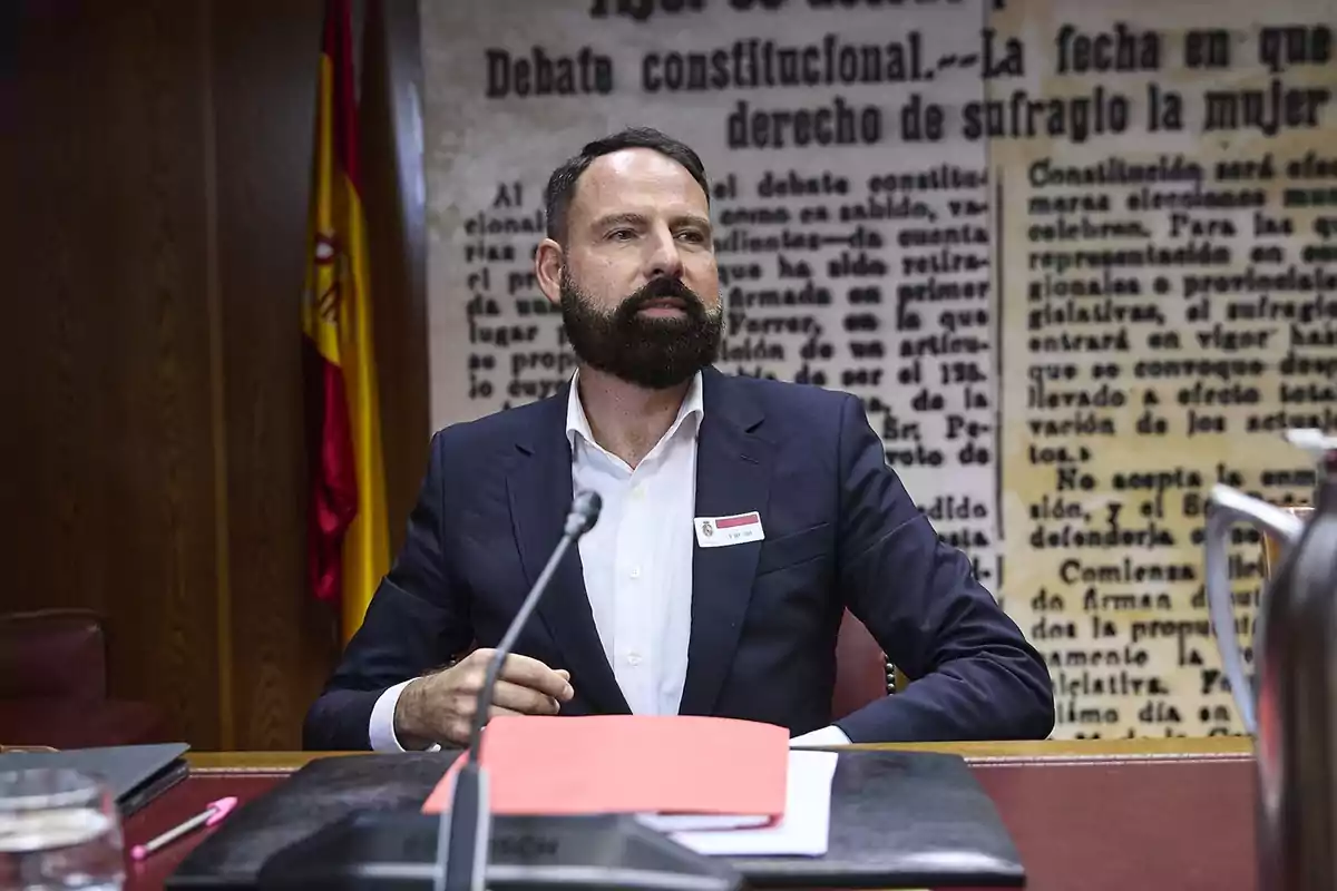 Hombre con barba y traje sentado en una mesa de trabajo con documentos y micrófono, al fondo una bandera de España y un mural con texto antiguo. Hombre con barba y traje sentado en una mesa de trabajo con documentos y micrófono, al fondo una bandera de España y un mural con texto antiguo.