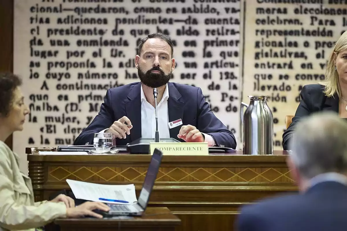Hombre con barba sentado en una mesa de comparecencia en una sala de reuniones, rodeado de otras personas y con fondo de texto grande en la pared Hombre con barba sentado en una mesa de comparecencia en una sala de reuniones, rodeado de otras personas y con fondo de texto grande en la pared