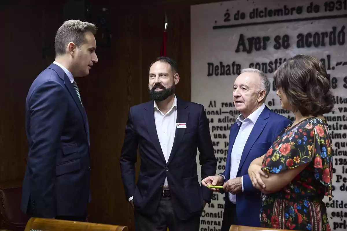 Cuatro personas conversan en una sala con paredes de madera y un mural con texto en el fondo Cuatro personas conversan en una sala con paredes de madera y un mural con texto en el fondo