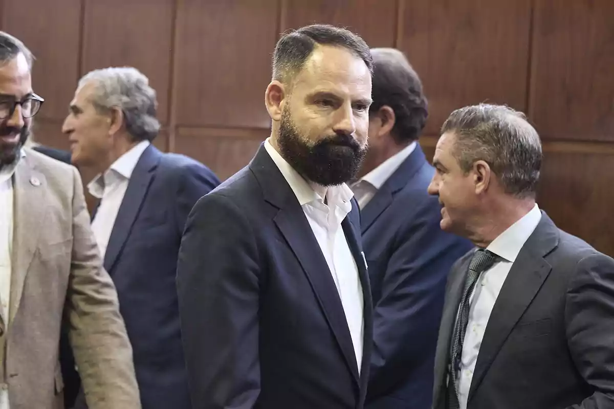 Un hombre de barba y traje oscuro está de pie en una sala con varias personas conversando a su alrededor