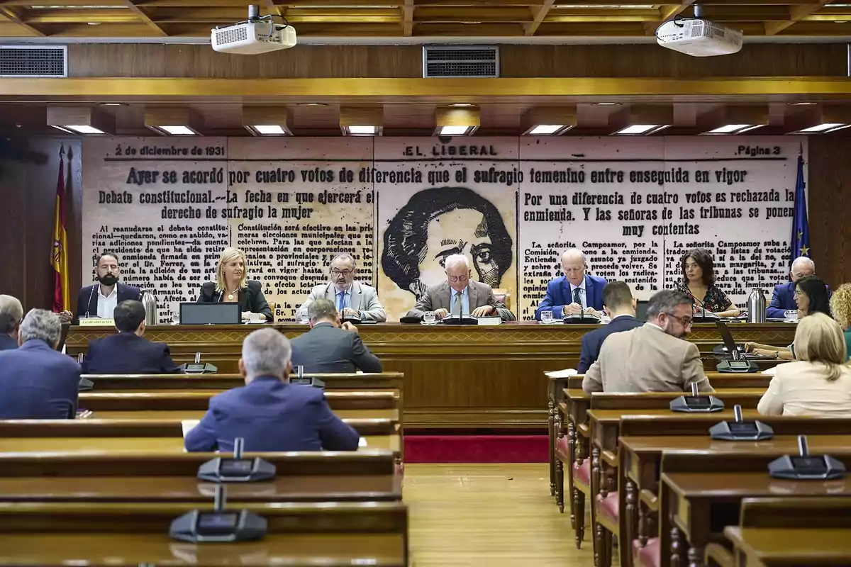 Personas sentadas en una sala de reuniones parlamentaria con un mural de fondo que muestra un retrato y un texto antiguo sobre el sufragio femenino en España Personas sentadas en una sala de reuniones parlamentaria con un mural de fondo que muestra un retrato y un texto antiguo sobre el sufragio femenino en España
