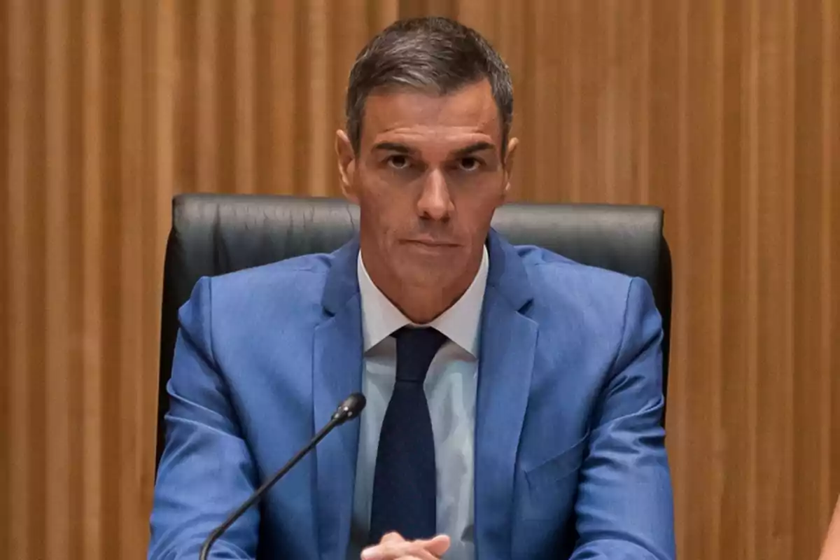 Hombre de cabello corto y canoso con traje azul sentado en una silla de respaldo alto frente a un micrófono en un entorno formal de madera