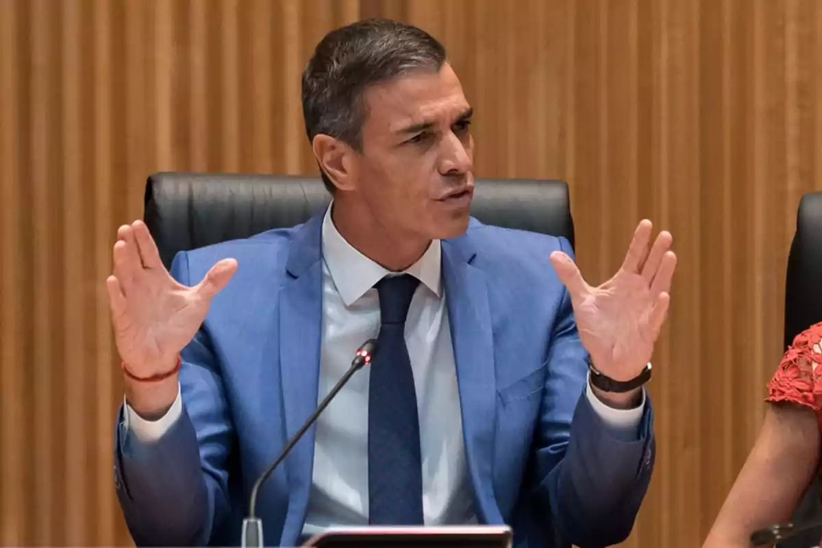Hombre de cabello corto y canoso con traje azul y corbata hablando en una reunión mientras gesticula con las manos.