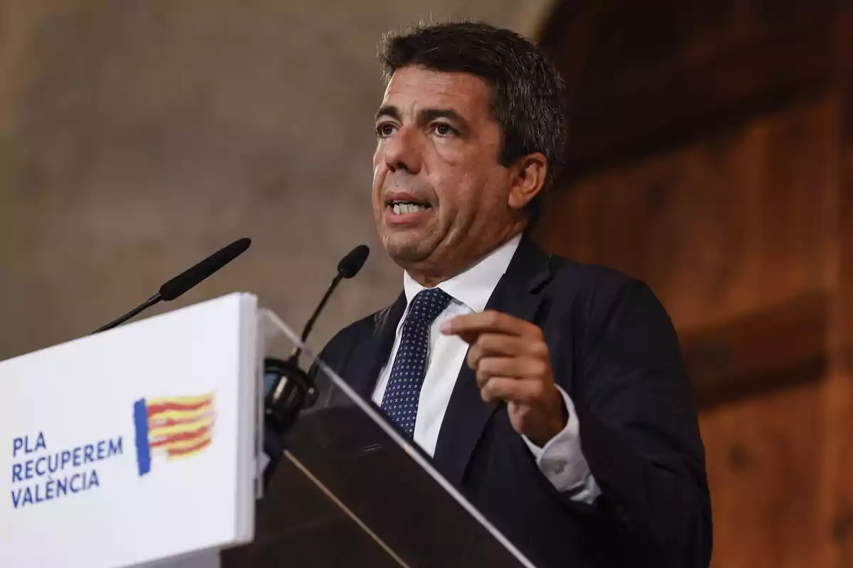 Hombre de traje hablando en un atril con micrófonos y un cartel que dice Pla Recuperem València Hombre de traje hablando en un atril con micrófonos y un cartel que dice Pla Recuperem València