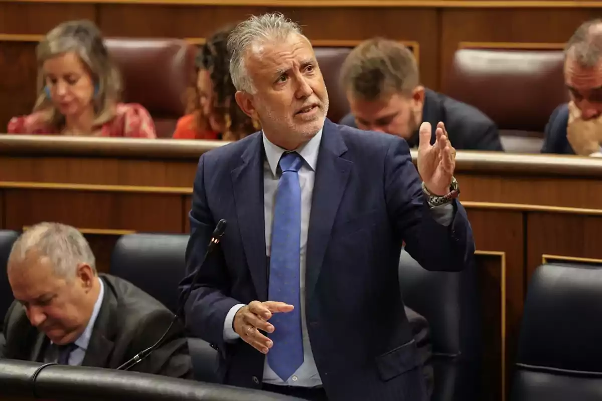 Hombre de cabello canoso y traje azul hablando en un parlamento mientras gesticula con la mano y otras personas están sentadas al fondo Hombre de cabello canoso y traje azul hablando en un parlamento mientras gesticula con la mano y otras personas están sentadas al fondo