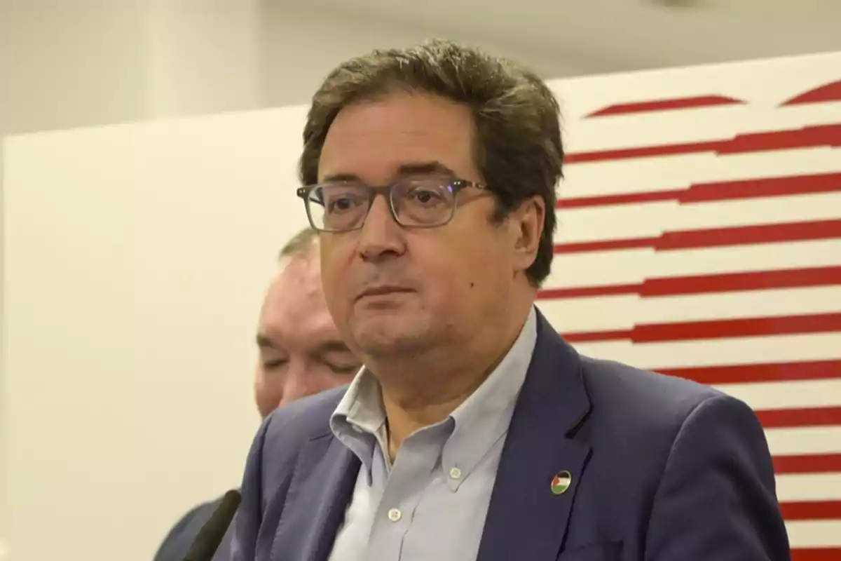 Hombre de cabello castaño y gafas con expresión seria usando saco azul y camisa clara frente a un fondo blanco con líneas rojas Hombre de cabello castaño y gafas con expresión seria usando saco azul y camisa clara frente a un fondo blanco con líneas rojas