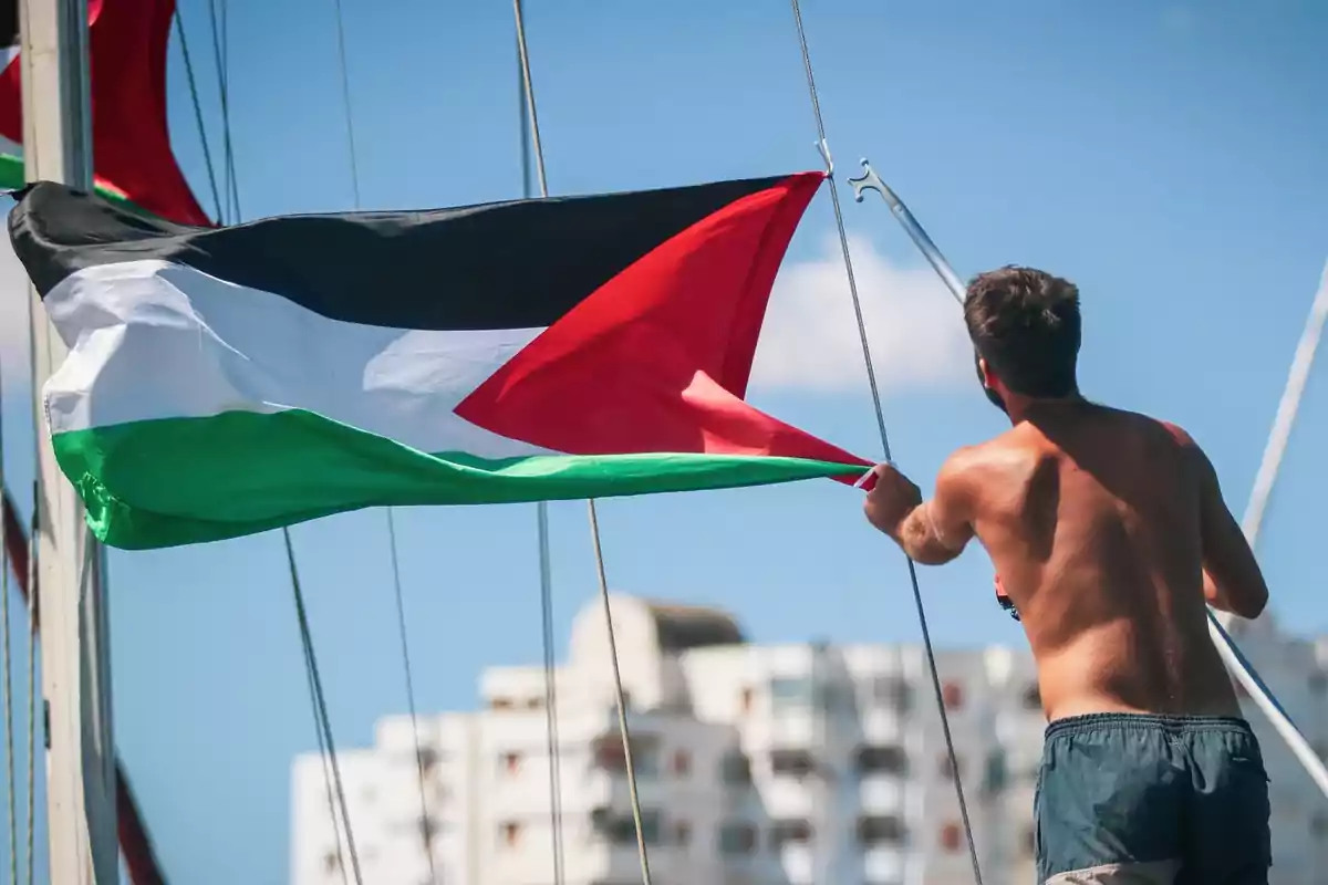 Hombre sin camiseta sostiene una bandera de Palestina en un barco con edificios al fondo y cielo despejado
