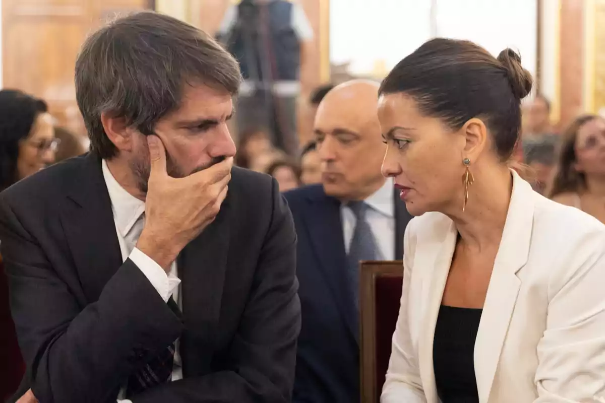 Dos personas vestidas de manera formal conversan de cerca en un evento con más asistentes al fondo Dos personas vestidas de manera formal conversan de cerca en un evento con más asistentes al fondo