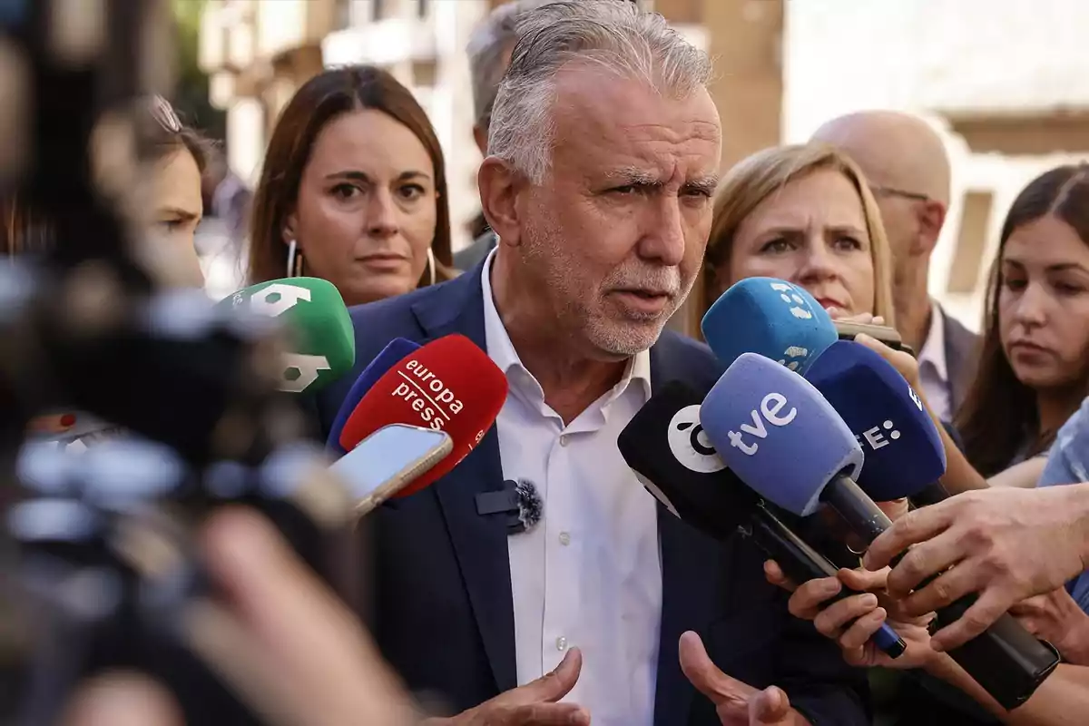 Un hombre de cabello canoso y traje oscuro responde preguntas de varios periodistas que sostienen micrófonos de diferentes medios de comunicación mientras varias personas lo observan atentamente Un hombre de cabello canoso y traje oscuro responde preguntas de varios periodistas que sostienen micrófonos de diferentes medios de comunicación mientras varias personas lo observan atentamente