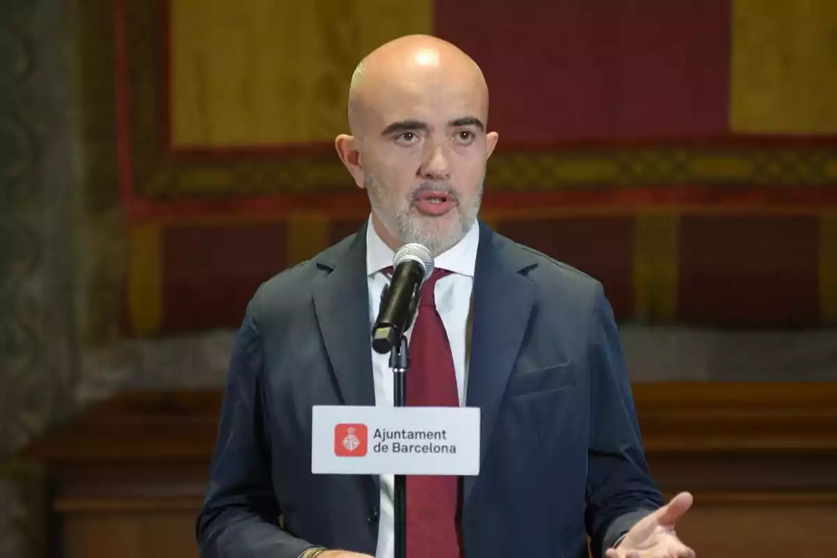 Hombre calvo con barba hablando frente a un micrófono con un cartel del Ayuntamiento de Barcelona en un entorno interior formal