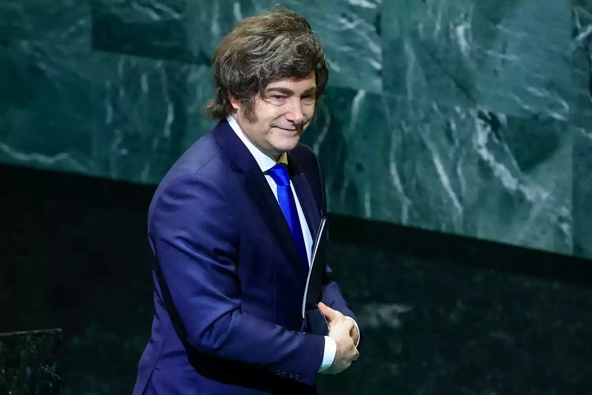 Hombre de cabello castaño y traje azul con corbata azul sonríe mientras sostiene unos papeles frente a una pared de mármol verde