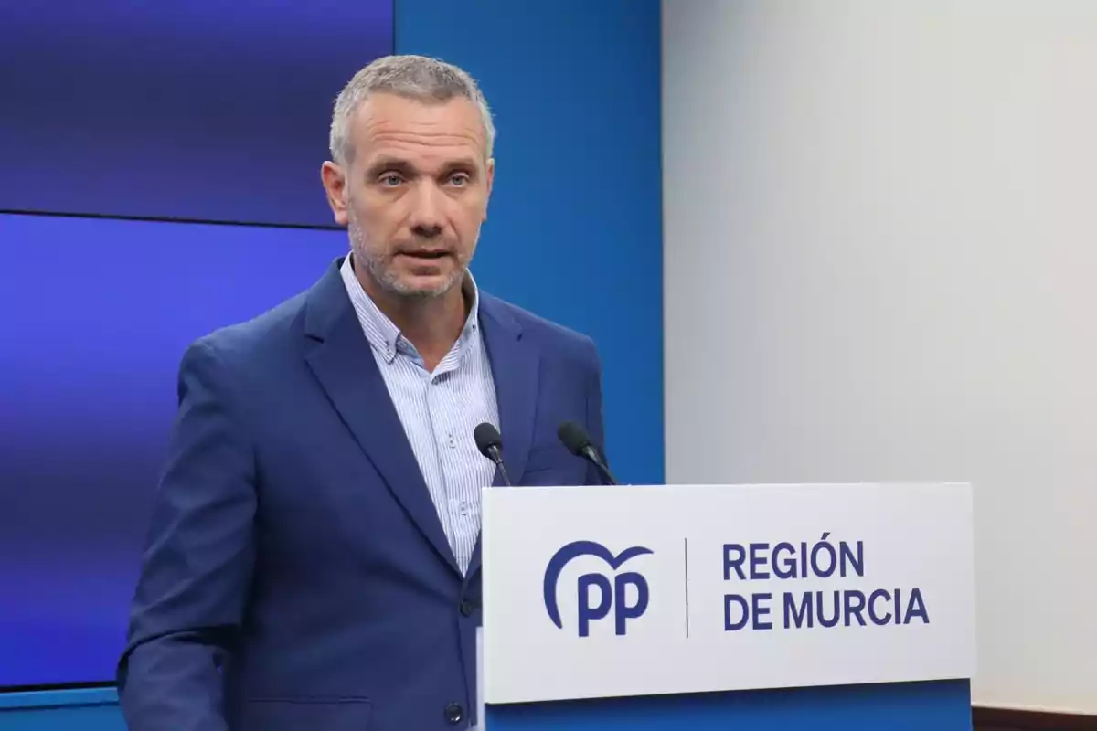 Hombre de cabello corto y canoso con barba, vestido con traje azul, hablando en un atril con el logo del PP y el texto Región de Murcia sobre un fondo azul y blanco Hombre de cabello corto y canoso con barba, vestido con traje azul, hablando en un atril con el logo del PP y el texto Región de Murcia sobre un fondo azul y blanco