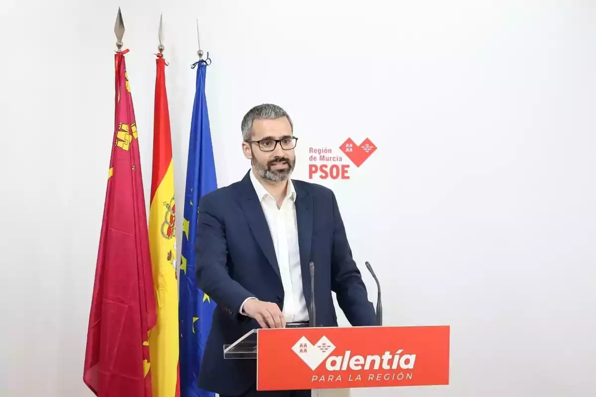Hombre con gafas y barba hablando en un atril del PSOE con banderas de fondo Hombre con gafas y barba hablando en un atril del PSOE con banderas de fondo