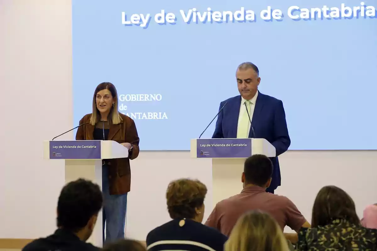 Dos personas en un escenario hablan frente a un público durante una presentación sobre la Ley de Vivienda de Cantabria con un fondo azul que muestra el título del evento. Dos personas en un escenario hablan frente a un público durante una presentación sobre la Ley de Vivienda de Cantabria con un fondo azul que muestra el título del evento.