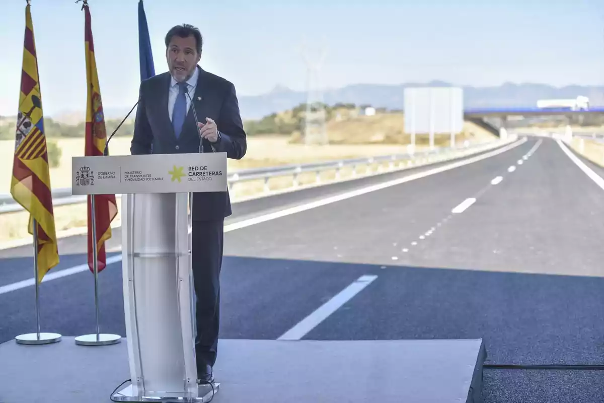 Un hombre con traje y corbata da un discurso en un podio sobre una carretera nueva, acompañado de banderas oficiales y un paisaje de fondo. Un hombre con traje y corbata da un discurso en un podio sobre una carretera nueva, acompañado de banderas oficiales y un paisaje de fondo.