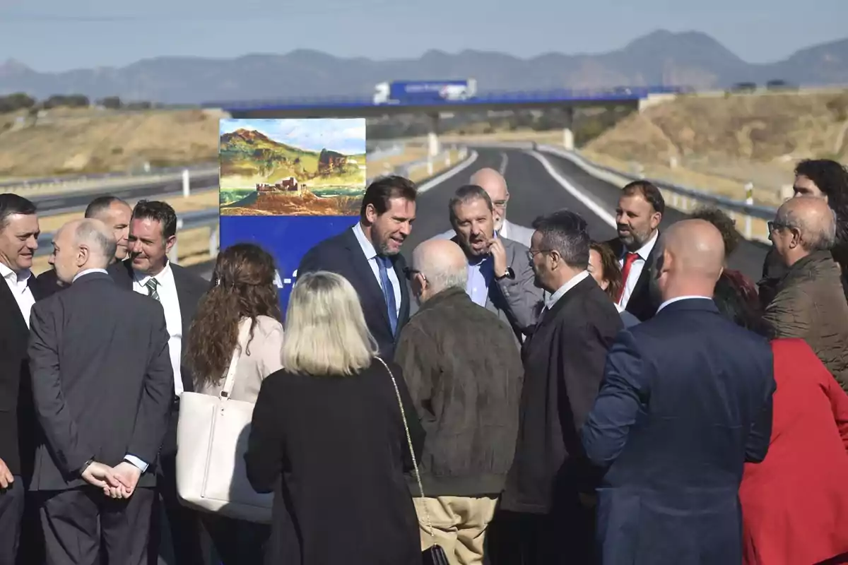 Un grupo de personas vestidas de manera formal conversa en medio de una carretera recién inaugurada con montañas al fondo y un cartel con una pintura paisajística Un grupo de personas vestidas de manera formal conversa en medio de una carretera recién inaugurada con montañas al fondo y un cartel con una pintura paisajística