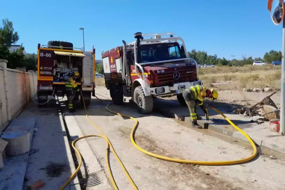 Dos camiones de bomberos y dos bomberos trabajando con mangueras en una zona al aire libre en un día soleado Dos camiones de bomberos y dos bomberos trabajando con mangueras en una zona al aire libre en un día soleado