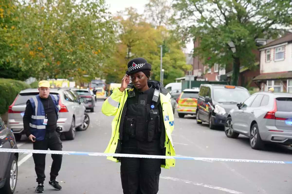 Una oficial de policía con chaqueta amarilla y uniforme negro está de pie detrás de una cinta de acordonamiento en una calle con varios autos y árboles, mientras otra persona con chaleco y gorra amarilla se encuentra al fondo. Una oficial de policía con chaqueta amarilla y uniforme negro está de pie detrás de una cinta de acordonamiento en una calle con varios autos y árboles, mientras otra persona con chaleco y gorra amarilla se encuentra al fondo.