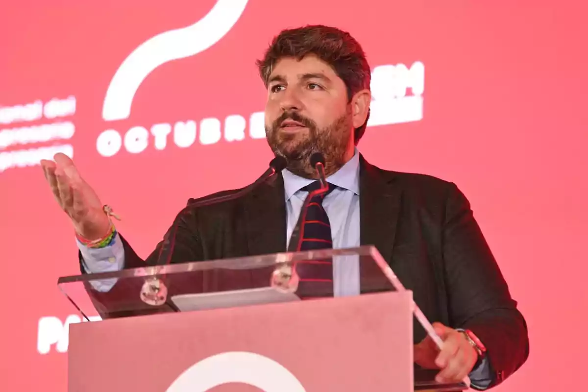 Hombre con barba y traje hablando en un podio frente a un fondo rojo durante un evento
