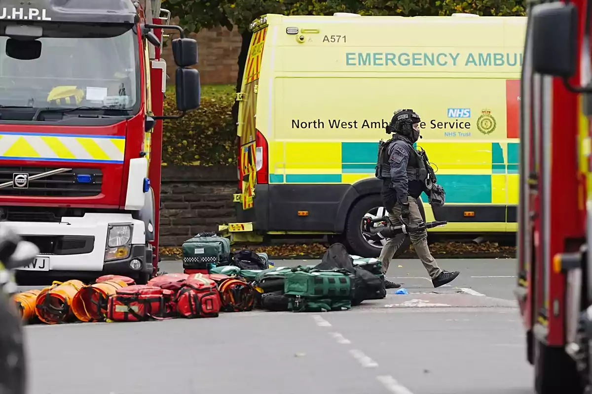 Un agente de seguridad camina entre equipos de emergencia frente a una ambulancia y un camión de bomberos en una calle Un agente de seguridad camina entre equipos de emergencia frente a una ambulancia y un camión de bomberos en una calle
