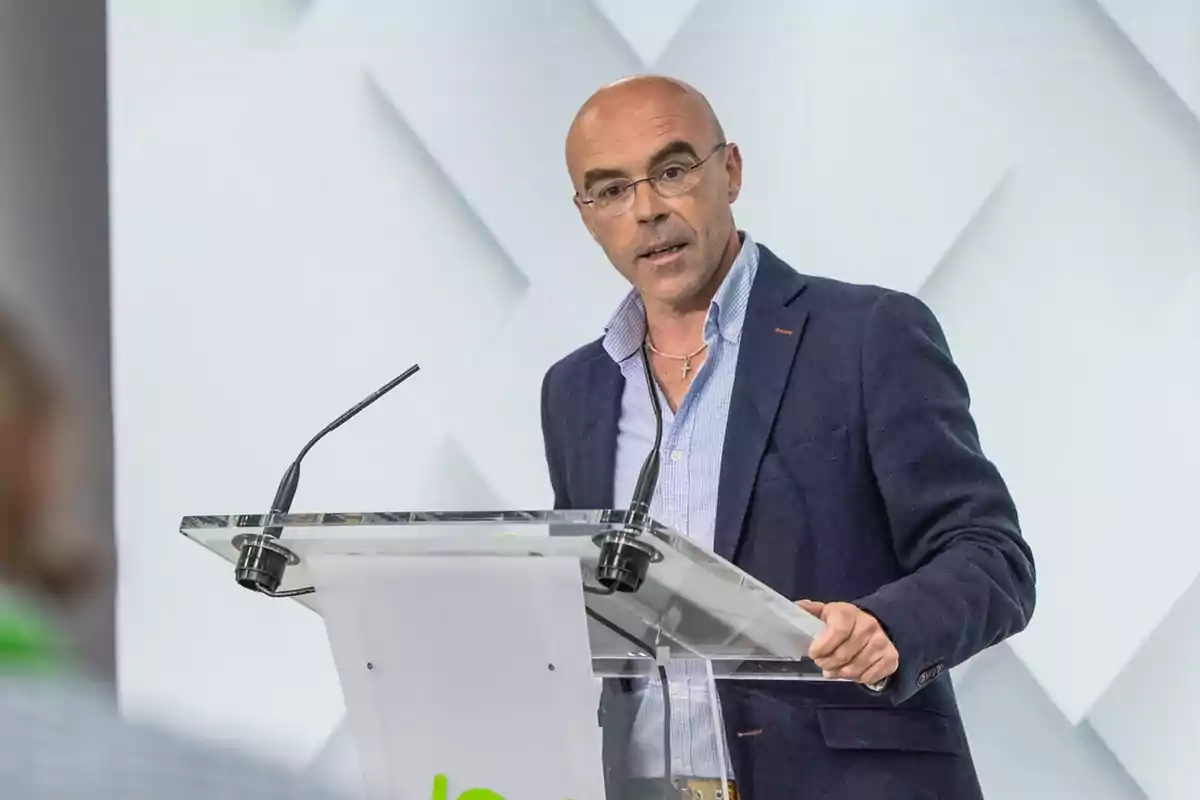 Hombre calvo con gafas y chaqueta azul hablando en un atril con micrófonos en un entorno profesional. Hombre calvo con gafas y chaqueta azul hablando en un atril con micrófonos en un entorno profesional.