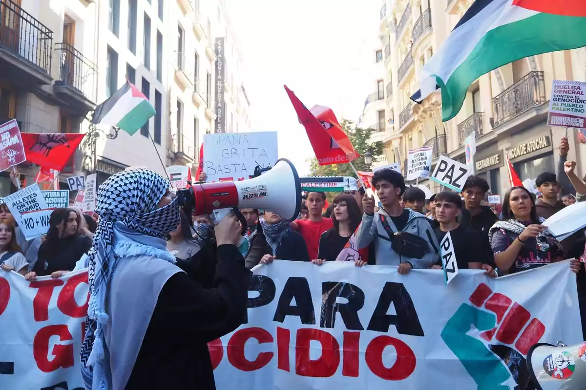Manifestación en una calle con personas sosteniendo pancartas y banderas en apoyo a Palestina, una persona con kufiya usa un megáfono mientras la multitud muestra mensajes de paz y protesta Manifestación en una calle con personas sosteniendo pancartas y banderas en apoyo a Palestina, una persona con kufiya usa un megáfono mientras la multitud muestra mensajes de paz y protesta