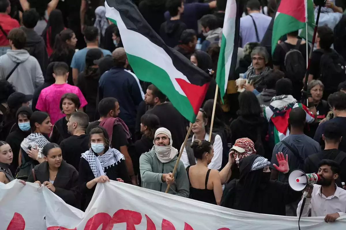 Manifestación con personas sosteniendo una pancarta y banderas palestinas entre una multitud