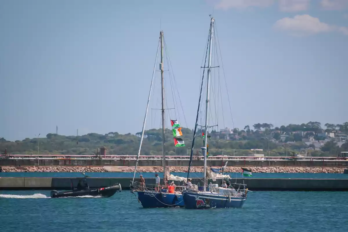Un velero azul con varias personas a bordo navega cerca de un muelle mientras una lancha de la policía pasa a su lado en el agua Un velero azul con varias personas a bordo navega cerca de un muelle mientras una lancha de la policía pasa a su lado en el agua