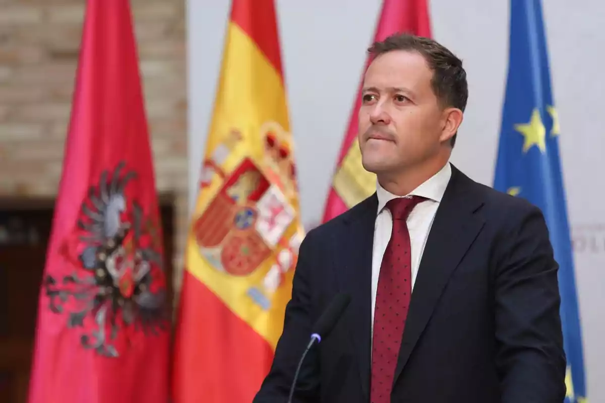 Hombre con traje oscuro y corbata roja de pie frente a micrófono con banderas oficiales de fondo