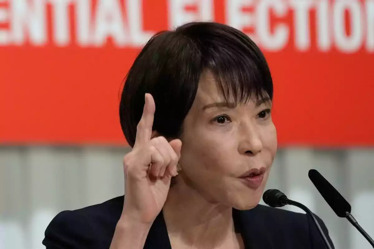 Mujer hablando en un micrófono con el dedo índice levantado frente a un fondo rojo con letras blancas borrosas Mujer hablando en un micrófono con el dedo índice levantado frente a un fondo rojo con letras blancas borrosas