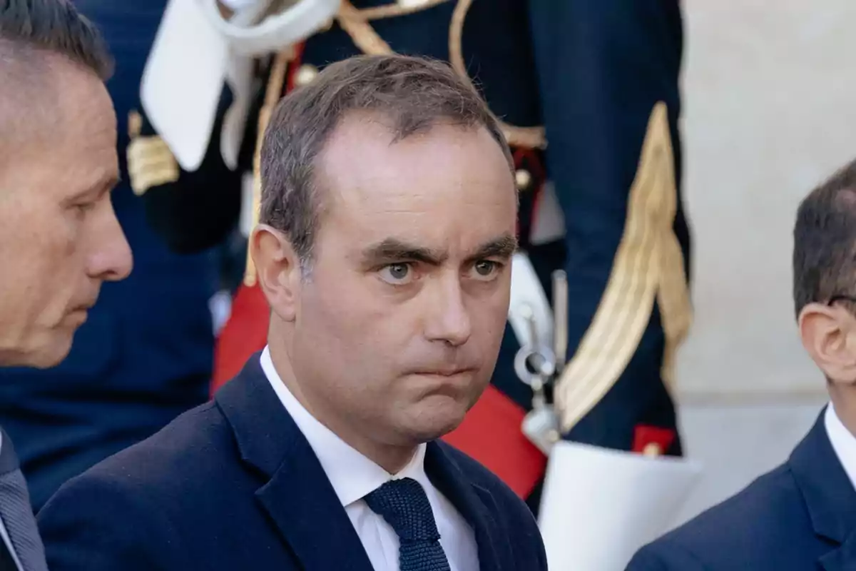 Hombre de traje azul con expresión seria rodeado de otras personas en un evento formal