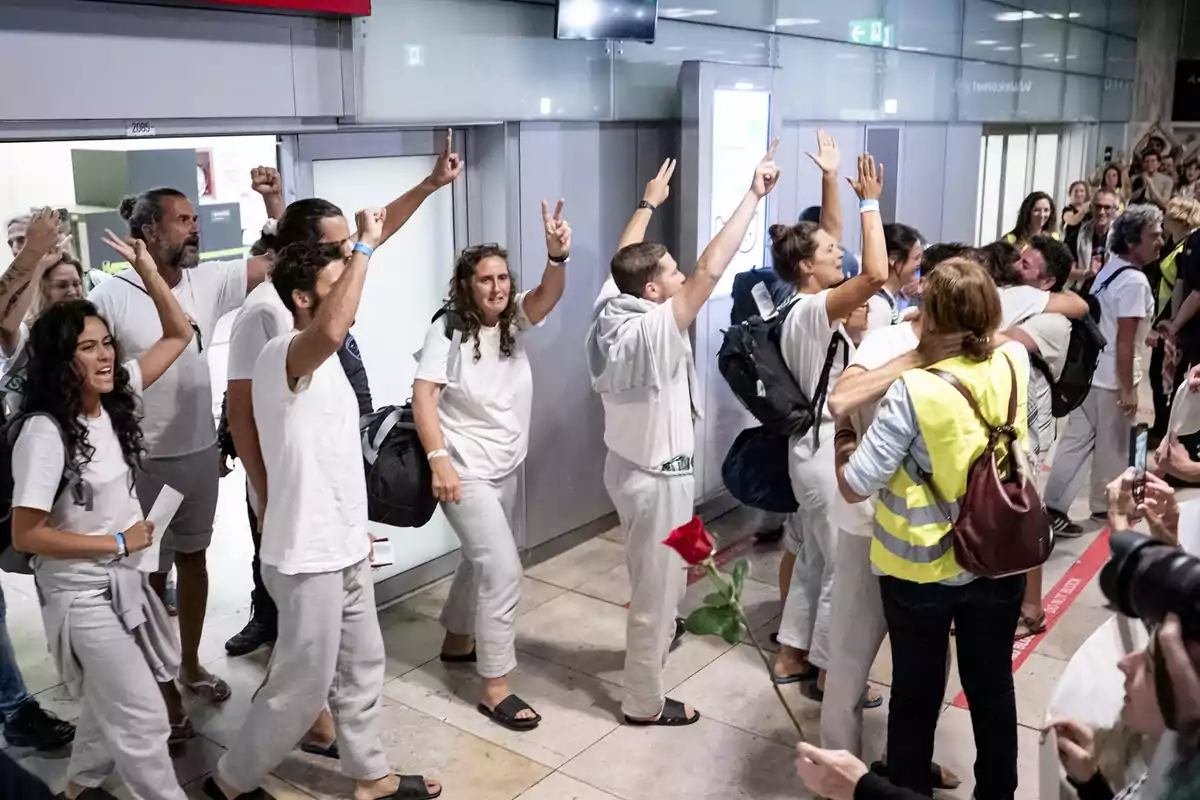 Un grupo de personas vestidas de blanco celebra y levanta los brazos al llegar a un aeropuerto mientras son recibidos por otras personas, una de ellas sostiene una rosa roja Un grupo de personas vestidas de blanco celebra y levanta los brazos al llegar a un aeropuerto mientras son recibidos por otras personas, una de ellas sostiene una rosa roja