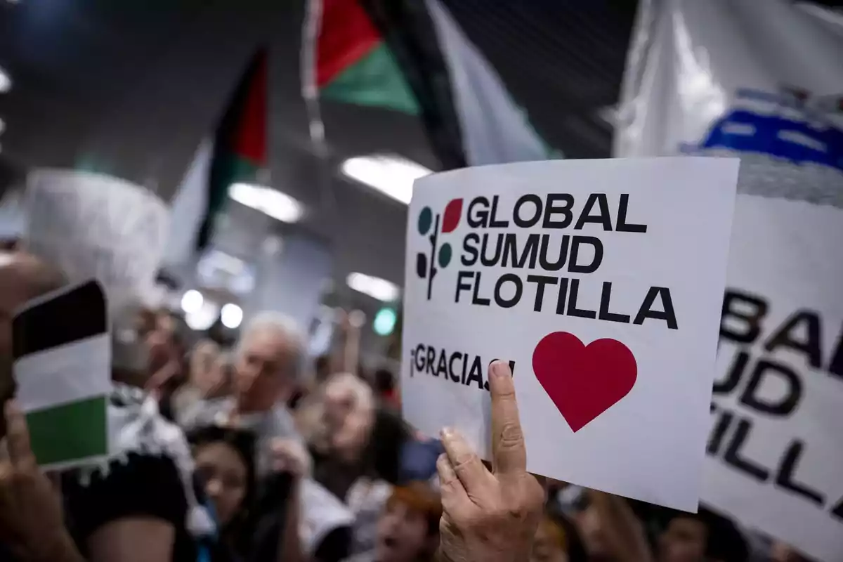 Persona sostiene un cartel que dice Global Sumud Flotilla y un corazón rojo en medio de una multitud con banderas y pancartas.
