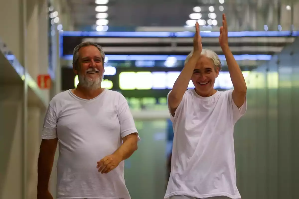 Dos personas sonrientes vestidas de blanco caminan por un pasillo iluminado mientras una de ellas aplaude con entusiasmo Dos personas sonrientes vestidas de blanco caminan por un pasillo iluminado mientras una de ellas aplaude con entusiasmo