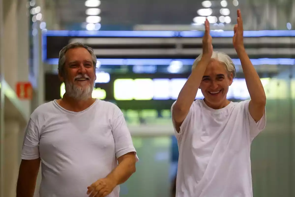 Dos personas sonríen y caminan juntas en un pasillo iluminado, una de ellas aplaude con entusiasmo