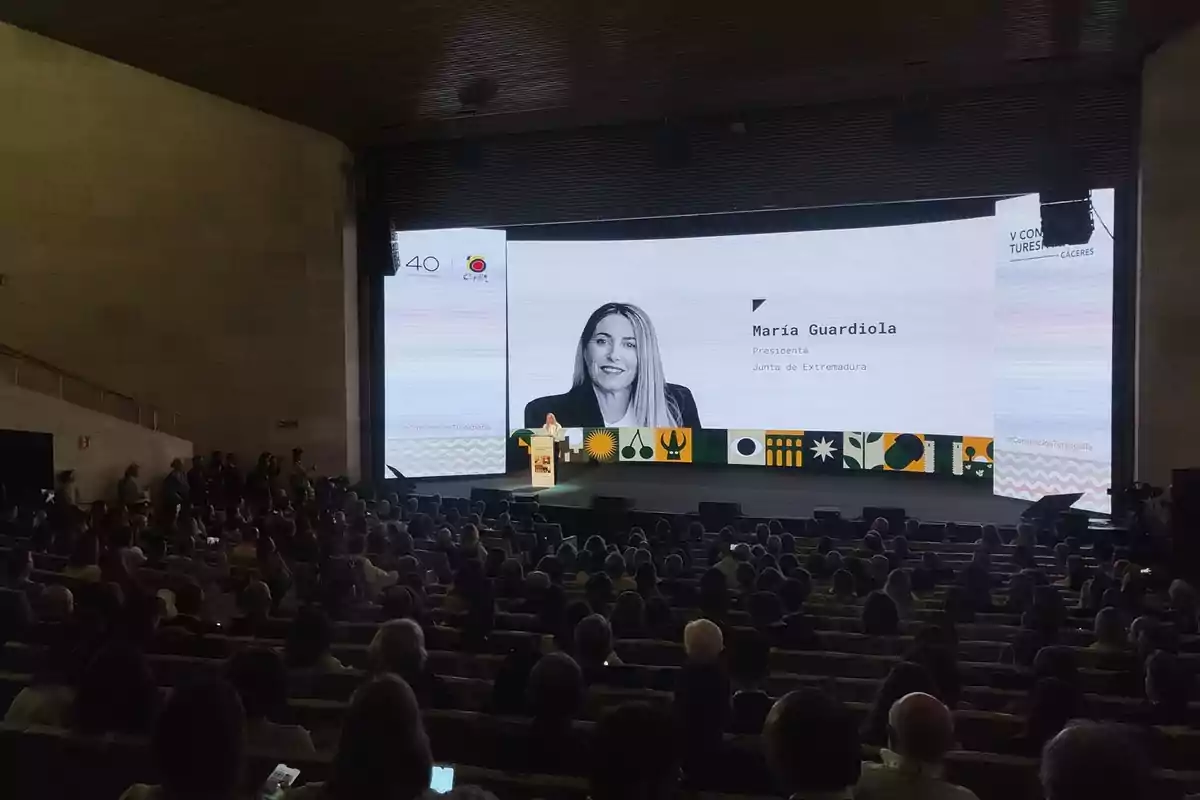 Auditorio lleno de personas asistiendo a una conferencia donde una mujer está dando un discurso en un escenario con una gran pantalla que muestra su imagen y su nombre Auditorio lleno de personas asistiendo a una conferencia donde una mujer está dando un discurso en un escenario con una gran pantalla que muestra su imagen y su nombre