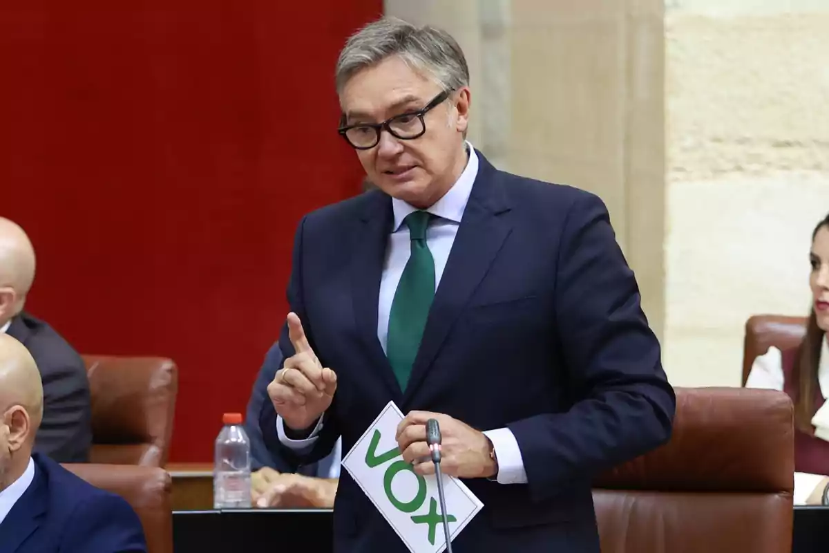 Hombre de traje y corbata verde hablando en un parlamento mientras sostiene un cartel con el logo de VOX Hombre de traje y corbata verde hablando en un parlamento mientras sostiene un cartel con el logo de VOX
