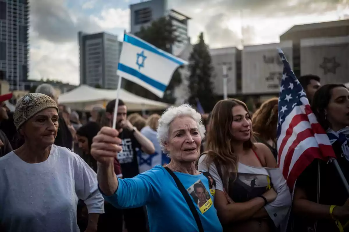 Mujer mayor sostiene una bandera de Israel en una manifestación junto a otras personas, una de ellas lleva una bandera de Estados Unidos, en un entorno urbano con edificios altos al fondo Mujer mayor sostiene una bandera de Israel en una manifestación junto a otras personas, una de ellas lleva una bandera de Estados Unidos, en un entorno urbano con edificios altos al fondo