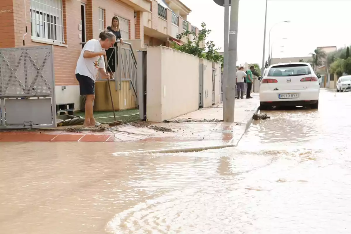 Una persona barre el lodo en la entrada de su casa mientras una calle está inundada y un coche blanco está estacionado cerca Una persona barre el lodo en la entrada de su casa mientras una calle está inundada y un coche blanco está estacionado cerca