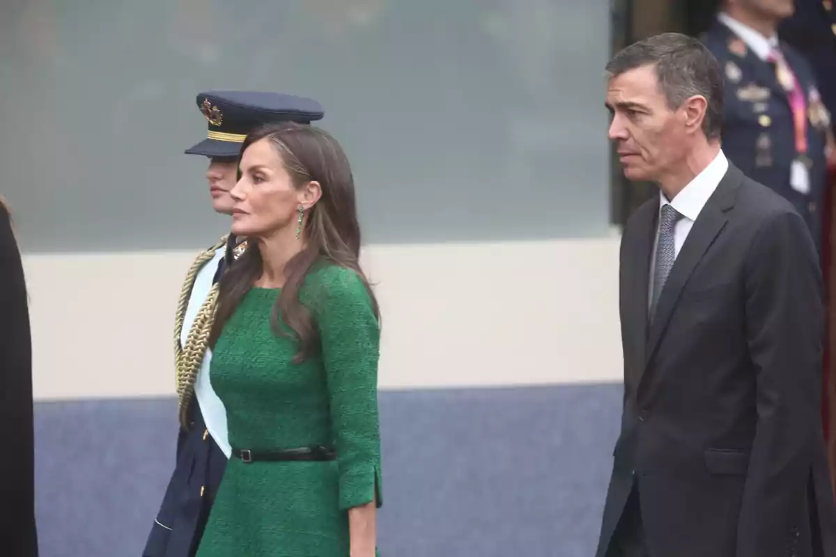 Una mujer con vestido verde y un hombre con traje oscuro caminan juntos en un evento formal, acompañados por una persona con uniforme militar.