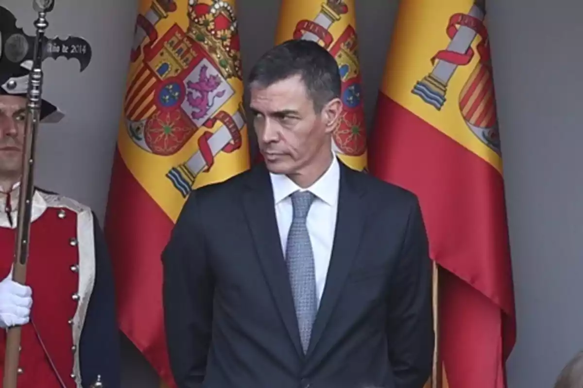 Hombre de traje oscuro y corbata gris de pie frente a varias banderas de España junto a un guardia con uniforme ceremonial