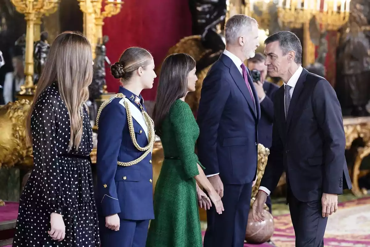 Cinco personas elegantemente vestidas se saludan en un salón decorado con detalles dorados y alfombra roja